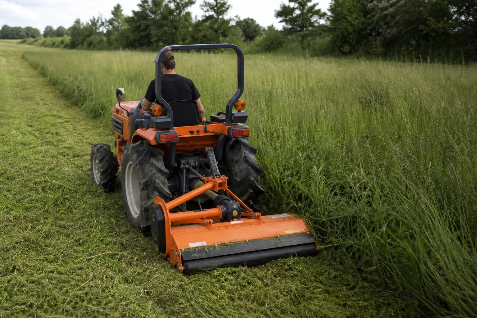 Malotraktor so zadným mulčovačom kosí pás vyššej trávy na vidieckom pozemku.