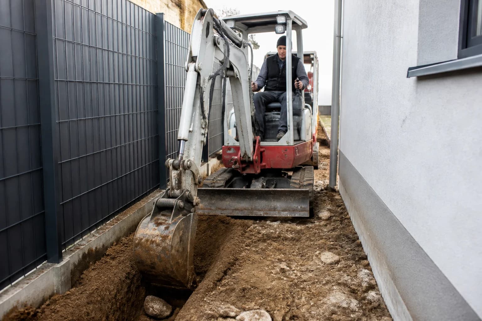 Kompaktný minibager pracuje v úzkom dvore medzi plotom a stenou domu.